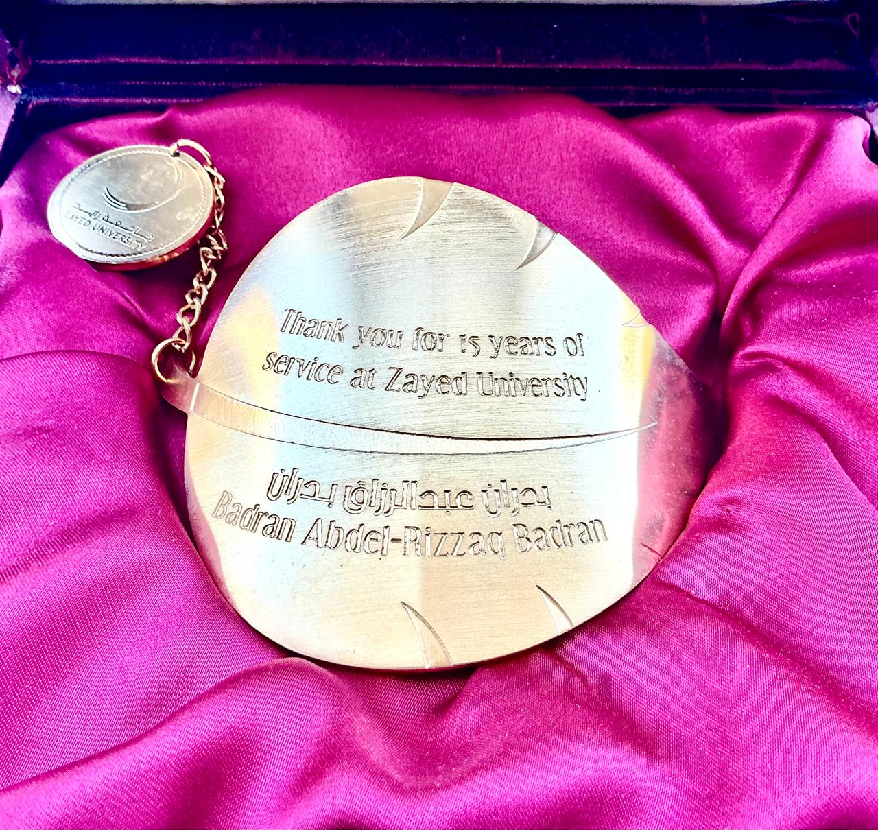 Gold medallion in a presentation box engraved: Thank you for 15 years of service at Zayed University, Badran Abdel-Rizzaq Badran, with a Zayed University seal on a chain